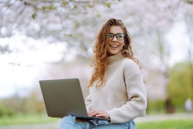 Gülen bir kadın lanet bir parkta dizüstü bilgisayarla masada oturuyor. Gözlük takan genç bir kadın gün batımında dışarıda dizüstü bilgisayarda çalışıyor. Uzaktan çalışma, rahatlama ve hafta sonları için kavramlar.