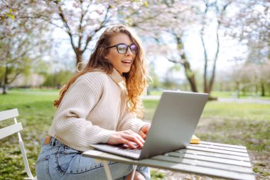 Gülen bir kadın lanet bir parkta dizüstü bilgisayarla masada oturuyor. Gözlük takan genç bir kadın gün batımında dışarıda dizüstü bilgisayarda çalışıyor. Uzaktan çalışma, rahatlama ve hafta sonları için kavramlar.