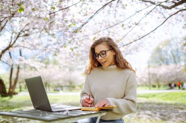 Gülen bir kadın lanet bir parkta dizüstü bilgisayarla masada oturuyor. Gözlük takan genç bir kadın gün batımında dışarıda dizüstü bilgisayarda çalışıyor. Uzaktan çalışma, rahatlama ve hafta sonları için kavramlar.