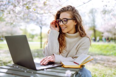 Gülen bir kadın lanet bir parkta dizüstü bilgisayarla masada oturuyor. Gözlük takan genç bir kadın gün batımında dışarıda dizüstü bilgisayarda çalışıyor. Uzaktan çalışma, rahatlama ve hafta sonları için kavramlar.
