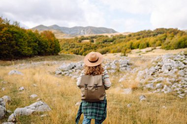 Arkadan bak. Sırt çantası ve şapkası olan bir yürüyüşçü kayalık dağ yolunda yürüyor. Genç kadın dağ manzarasının tadını çıkarıyor ve özgürlük duygusunu hissediyor. Yürüyüş, doğa ve seyahat kavramı.