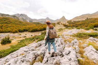 Arkadan bak. Sırt çantası ve şapkası olan bir yürüyüşçü kayalık dağ yolunda yürüyor. Genç kadın dağ manzarasının tadını çıkarıyor ve özgürlük duygusunu hissediyor. Yürüyüş, doğa ve seyahat kavramı.