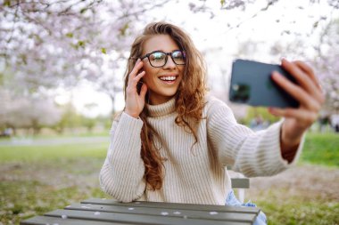 Güneşli bir günde çiçek açan bir parkta selfie çeken genç bir kadının portresi. Mutlu bir kadın gün batımında telefonuyla dışarıda blog yazıyor. Blog yazma, rahatlama ve hafta sonları kavramları.