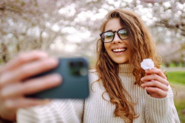 Güneşli bir günde çiçek açan bir parkta selfie çeken genç bir kadının portresi. Mutlu bir kadın gün batımında telefonuyla dışarıda blog yazıyor. Blog yazma, rahatlama ve hafta sonları kavramları.
