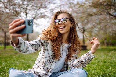 Portrait of a young woman taking a selfie in a blooming park on a sunny day. A happy woman blogs on her phone outdoors at sunset. Concepts of blogging, relaxation, and weekends.