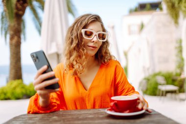 Genç bir kadın, elinde telefonla açık hava kafenin terasında güneşli bir günün tadını çıkarıyor. Zarif bir kadın palmiye ağaçlarıyla çevrili masada otururken telefonda kahve içip mesajlaşır. Tarz ve moda kavramı.