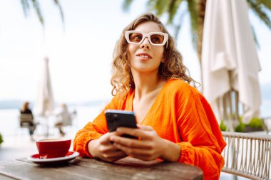 Genç bir kadın, elinde telefonla açık hava kafenin terasında güneşli bir günün tadını çıkarıyor. Zarif bir kadın palmiye ağaçlarıyla çevrili masada otururken telefonda kahve içip mesajlaşır. Tarz ve moda kavramı.