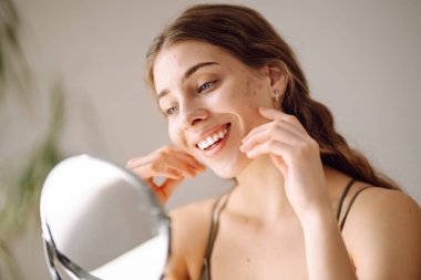 Genç bir kadının küçük aynadaki yansımasına ve güneşli odada tenine dokunuşuna yakından bakın. Güzel bir kadın sıcak bir odada masada otururken patlağı inceliyor. Sivilce kavramı, doğal güzellik.