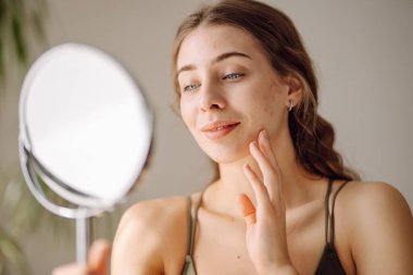 Genç bir kadının küçük aynadaki yansımasına ve güneşli odada tenine dokunuşuna yakından bakın. Güzel bir kadın sıcak bir odada masada otururken patlağı inceliyor. Sivilce kavramı, doğal güzellik.