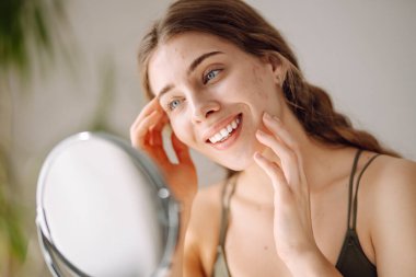 Genç bir kadının küçük aynadaki yansımasına ve güneşli odada tenine dokunuşuna yakından bakın. Güzel bir kadın sıcak bir odada masada otururken patlağı inceliyor. Sivilce kavramı, doğal güzellik.