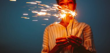 Koyu mavi bir gökyüzünün altında elinde maytap tutan bir kadın. O kutlama yaparken, kıvılcımlar parlak kıvılcımlar oluşturuyor. Bu sahne bir sevinç ve heyecan anı yakalar..