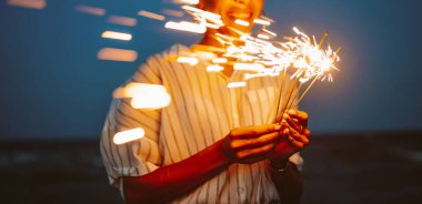 Koyu mavi bir gökyüzünün altında elinde maytap tutan bir kadın. O kutlama yaparken, kıvılcımlar parlak kıvılcımlar oluşturuyor. Bu sahne bir sevinç ve heyecan anı yakalar..