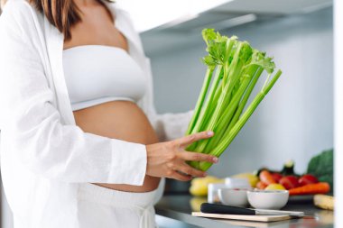 Hamile bir kadın, güneşli bir günde modern bir mutfakta kereviz ve diğer sebzeleri doğruyor. Hamileliğinin sonlarına doğru genç bir kadın sıcak bir mutfakta yemek hazırlıyor. Konseptler: sağlık, beklenti, yaşam tarzı.