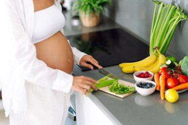 Hamile bir kadın, güneşli bir günde modern bir mutfakta kereviz ve diğer sebzeleri doğruyor. Hamileliğinin sonlarına doğru genç bir kadın sıcak bir mutfakta yemek hazırlıyor. Konseptler: sağlık, beklenti, yaşam tarzı.