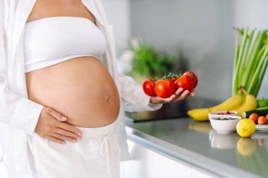 Hamile bir kadın, güneşli bir günde modern bir mutfakta kereviz ve diğer sebzeleri doğruyor. Hamileliğinin sonlarına doğru genç bir kadın sıcak bir mutfakta yemek hazırlıyor. Konseptler: sağlık, beklenti, yaşam tarzı.