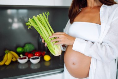 Hamile bir kadın, güneşli bir günde modern bir mutfakta kereviz ve diğer sebzeleri doğruyor. Hamileliğinin sonlarına doğru genç bir kadın sıcak bir mutfakta yemek hazırlıyor. Konseptler: sağlık, beklenti, yaşam tarzı.