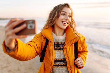 Sırt çantalı biri güneş ışınlarıyla sahilde selfie çeker. Genç bir kadın açık havada blog yazarak deniz kenarında vakit geçirmekten hoşlanıyor. Blog yazma, gevşeme ve hafta sonları için kavramlar.