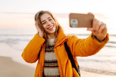 Sırt çantalı biri güneş ışınlarıyla sahilde selfie çeker. Genç bir kadın açık havada blog yazarak deniz kenarında vakit geçirmekten hoşlanıyor. Blog yazma, gevşeme ve hafta sonları için kavramlar.