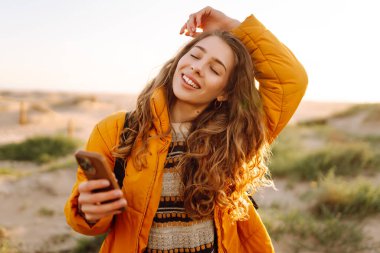 Güzel bir kadın sahilde duruyor, gün ışığında telefona bakıyor. Yürüyüş yolunda sırt çantası ve telefonu olan genç bir kadın deniz manzarasından hoşlanıyor. Teknoloji, rahatlama ve macera kavramı.