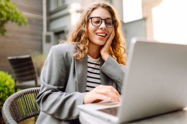 Güzel bir iş kadını dışarıda bir kafede dizüstü bilgisayarla oturuyor. Genç bir kadın dizüstü bilgisayarda çalışıyor ve terasta güneşli bir günün tadını çıkarıyor. Kavram: Uzaktan çalışma, rahatlama. Yaşam biçimi.