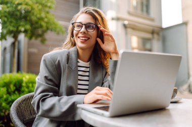 Güzel bir iş kadını dışarıda bir kafede dizüstü bilgisayarla oturuyor. Genç bir kadın dizüstü bilgisayarda çalışıyor ve terasta güneşli bir günün tadını çıkarıyor. Kavram: Uzaktan çalışma, rahatlama. Yaşam biçimi.