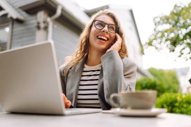 Güzel bir iş kadını dışarıda bir kafede dizüstü bilgisayarla oturuyor. Genç bir kadın dizüstü bilgisayarda çalışıyor ve terasta güneşli bir günün tadını çıkarıyor. Kavram: Uzaktan çalışma, rahatlama. Yaşam biçimi.