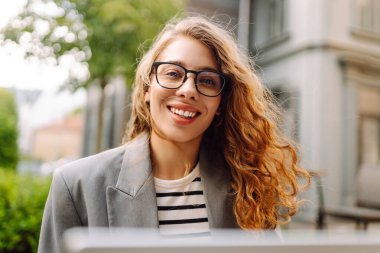 Güzel bir iş kadını dışarıda bir kafede dizüstü bilgisayarla oturuyor. Genç bir kadın dizüstü bilgisayarda çalışıyor ve terasta güneşli bir günün tadını çıkarıyor. Kavram: Uzaktan çalışma, rahatlama. Yaşam biçimi.