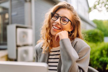 Güzel bir iş kadını dışarıda bir kafede dizüstü bilgisayarla oturuyor. Genç bir kadın dizüstü bilgisayarda çalışıyor ve terasta güneşli bir günün tadını çıkarıyor. Kavram: Uzaktan çalışma, rahatlama. Yaşam biçimi.