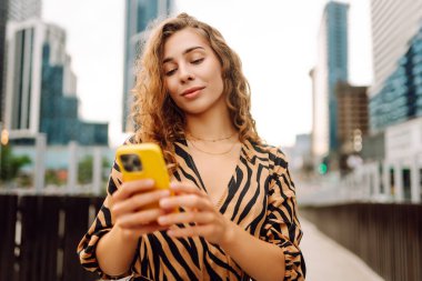 Şehirdeki metropol manzaralı bir caddede telefonu olan mutlu bir kadının portresi. Genç bir turist mesaj atıyor ve telefonuyla gün batımında gezintiye çıkıyor. Teknoloji, şehir ve hafta sonu kavramları.