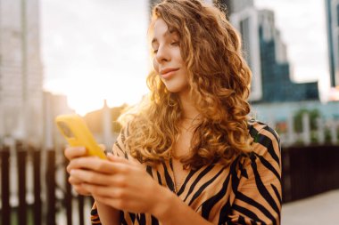 Şehirdeki metropol manzaralı bir caddede telefonu olan mutlu bir kadının portresi. Genç bir turist mesaj atıyor ve telefonuyla gün batımında gezintiye çıkıyor. Teknoloji, şehir ve hafta sonu kavramları.