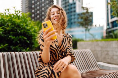 Şehirdeki metropol manzaralı bir caddede telefonu olan mutlu bir kadının portresi. Genç bir turist mesaj atıyor ve telefonuyla gün batımında gezintiye çıkıyor. Teknoloji, şehir ve hafta sonu kavramları.