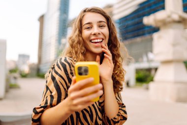 Şehirdeki metropol manzaralı bir caddede telefonu olan mutlu bir kadının portresi. Genç bir turist mesaj atıyor ve telefonuyla gün batımında gezintiye çıkıyor. Teknoloji, şehir ve hafta sonu kavramları.