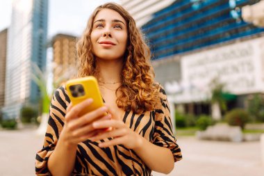Şehirdeki metropol manzaralı bir caddede telefonu olan mutlu bir kadının portresi. Genç bir turist mesaj atıyor ve telefonuyla gün batımında gezintiye çıkıyor. Teknoloji, şehir ve hafta sonu kavramları.