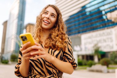 Şehirdeki metropol manzaralı bir caddede telefonu olan mutlu bir kadının portresi. Genç bir turist mesaj atıyor ve telefonuyla gün batımında gezintiye çıkıyor. Teknoloji, şehir ve hafta sonu kavramları.