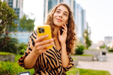 Şehirdeki metropol manzaralı bir caddede telefonu olan mutlu bir kadının portresi. Genç bir turist mesaj atıyor ve telefonuyla gün batımında gezintiye çıkıyor. Teknoloji, şehir ve hafta sonu kavramları.