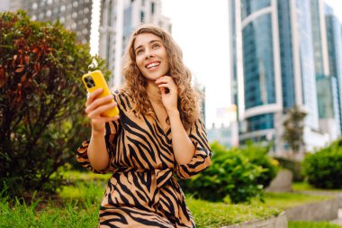 Şehirdeki metropol manzaralı bir caddede telefonu olan mutlu bir kadının portresi. Genç bir turist mesaj atıyor ve telefonuyla gün batımında gezintiye çıkıyor. Teknoloji, şehir ve hafta sonu kavramları.