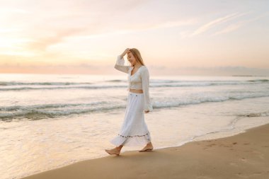 Genç bir kadın güneşli bir sahilde yürüyor, deniz manzarasının tadını çıkarıyor. Güzel bir kadın kendini özgür hisseder ve gün batımına hayran kalır. Kavram: rahatlama, doğa.