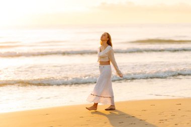 Genç bir kadın güneşli bir sahilde yürüyor, deniz manzarasının tadını çıkarıyor. Güzel bir kadın kendini özgür hisseder ve gün batımına hayran kalır. Kavram: rahatlama, doğa.