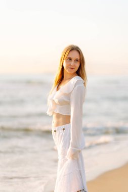 Genç bir kadın güneşli bir sahilde yürüyor, deniz manzarasının tadını çıkarıyor. Güzel bir kadın kendini özgür hisseder ve gün batımına hayran kalır. Kavram: rahatlama, doğa.