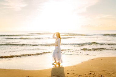 Genç bir kadın güneşli bir sahilde yürüyor, deniz manzarasının tadını çıkarıyor. Güzel bir kadın kendini özgür hisseder ve gün batımına hayran kalır. Kavram: rahatlama, doğa.