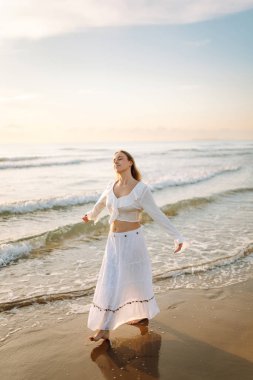 Genç bir kadın güneşli bir sahilde yürüyor, deniz manzarasının tadını çıkarıyor. Güzel bir kadın kendini özgür hisseder ve gün batımına hayran kalır. Kavram: rahatlama, doğa.