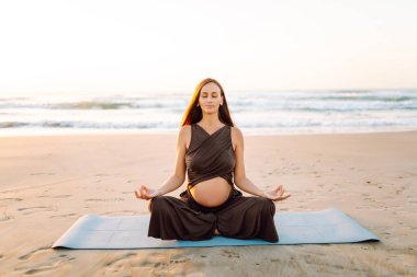 Spor elbiseli hamile bir kadın gün batımında plajda yoga yapıyor. Müstakbel anne deniz manzarasını sever ve dışarıda meditasyon yapar. Hamilelik, rahatlama ve sağlık kavramları.