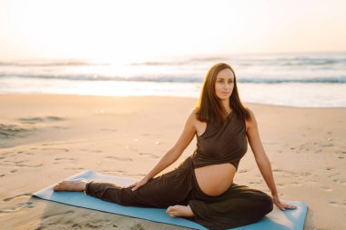 Spor elbiseli hamile bir kadın gün batımında plajda yoga yapıyor. Müstakbel anne deniz manzarasını sever ve dışarıda meditasyon yapar. Hamilelik, rahatlama ve sağlık kavramları.