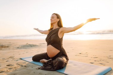 Spor elbiseli hamile bir kadın gün batımında plajda yoga yapıyor. Müstakbel anne deniz manzarasını sever ve dışarıda meditasyon yapar. Hamilelik, rahatlama ve sağlık kavramları.