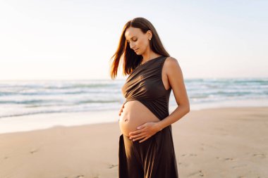 Spor elbiseli hamile bir kadın gün batımında plajda yoga yapıyor. Müstakbel anne deniz manzarasını sever ve dışarıda meditasyon yapar. Hamilelik, rahatlama ve sağlık kavramları.