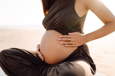 Spor elbiseli hamile bir kadın gün batımında plajda yoga yapıyor. Müstakbel anne deniz manzarasını sever ve dışarıda meditasyon yapar. Hamilelik, rahatlama ve sağlık kavramları.