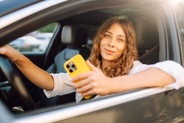 Kıvırcık saçlı ve telefonu olan genç bir kadın arabanın direksiyonunda oturur ve güneşli bir günde mesajlaşır. Güzel bir kadın elinde telefonla gezmekten zevk alır. Kentsel ulaşım kavramı.