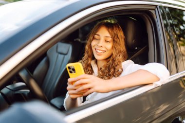 Kıvırcık saçlı ve telefonu olan genç bir kadın arabanın direksiyonunda oturur ve güneşli bir günde mesajlaşır. Güzel bir kadın elinde telefonla gezmekten zevk alır. Kentsel ulaşım kavramı.