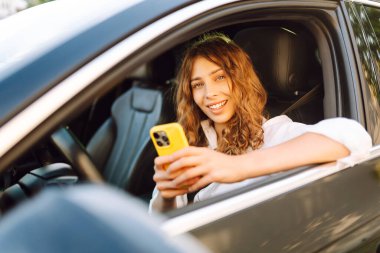 Kıvırcık saçlı ve telefonu olan genç bir kadın arabanın direksiyonunda oturur ve güneşli bir günde mesajlaşır. Güzel bir kadın elinde telefonla gezmekten zevk alır. Kentsel ulaşım kavramı.