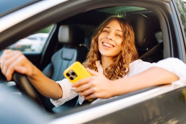 Kıvırcık saçlı ve telefonu olan genç bir kadın arabanın direksiyonunda oturur ve güneşli bir günde mesajlaşır. Güzel bir kadın elinde telefonla gezmekten zevk alır. Kentsel ulaşım kavramı.